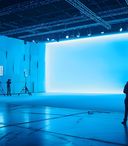 A large professional film or photography studio filled with equipment and crew members. The space is dominated by a huge, seamless bright blue backdrop that curves at the base, creating an infinity‑style stage. Several silhouetted figures stand or work around cameras, tripods, and large LED panel lights positioned throughout the room. The floor reflects the blue light, and overhead rigging and studio fixtures are visible across the dark ceiling.