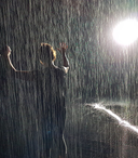 Photo of person holding hands up as if dancing in the rain with what looks like rain and a bright spotlight behind them 