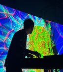 an image showing a silhouette of a man using a laptop and mixing desk with a colourful computer generated graphic showing on large screen as a backdrop