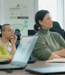 Four women of diverse backgrounds sit at a table in a bright classroom, attentively listening during a workshop, with laptops, notebooks, a coffee cup, and a water bottle on the table.