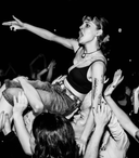 Black-and-white photo of a female crowd-surfing performer being carried above a packed concert crowd. She wears a black crop top and jeans, has tattoos on her left arm, and points outward with her right arm while fans reach up to support her