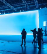 A large professional film or photography studio filled with equipment and crew members. The space is dominated by a huge, seamless bright blue backdrop that curves at the base, creating an infinity‑style stage. Several silhouetted figures stand or work around cameras, tripods, and large LED panel lights positioned throughout the room. The floor reflects the blue light, and overhead rigging and studio fixtures are visible across the dark ceiling.