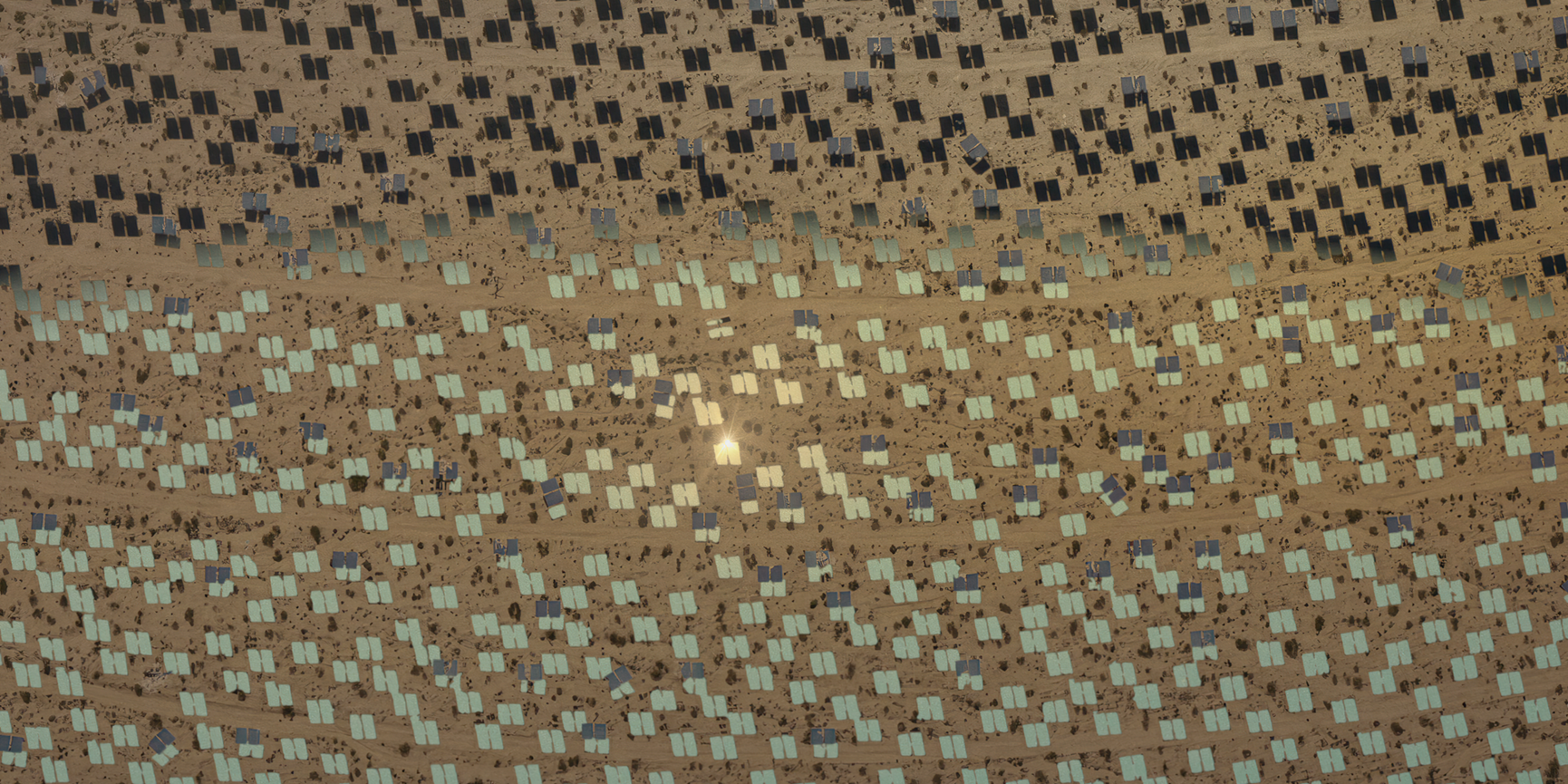 A film still from The Great Endeavour, directed and designed by Liam Young, 2024. The image shows an aerial view of a large solar farm in a desert landscape. Rows of solar panels are arranged in a grid pattern, with some sections appearing darker and others lighter, possibly due to the angle of sunlight or different types of panels. The ground is sandy and dotted with sparse vegetation. There is a bright reflection of sunlight near the centre of the image.
