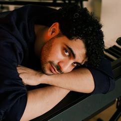 Portrait of a man with brown skin and dark hair leaning on a keyboard with his head on his forearm looking sideways at the camera
