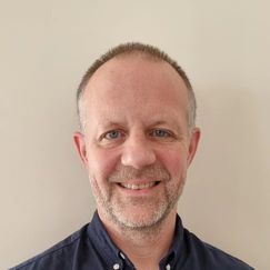 A person wearing a dark button‑up shirt stands against a plain light‑coloured wall.