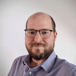 A person in a light purple collared shirt stands against a plain light background.