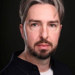 A person shown from the shoulders up, wearing a dark button-up shirt over a light-colored top, standing against a dark background.
