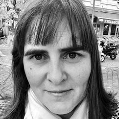 A person with medium-length hair stands outdoors in a black‑and‑white photo, with shops and parked scooters visible in the background.