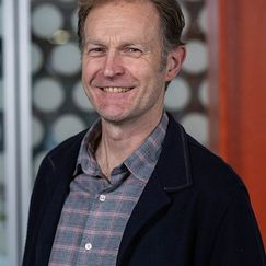 A person wearing a plaid shirt and a dark jacket stands indoors in front of a patterned background.