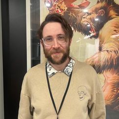 A person wearing a beige sweater with a skull design stands in front of a large poster featuring a stylized creature.