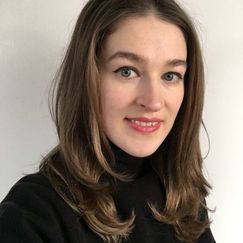 A person with long hair that includes a blue streak is wearing a dark jacket and light top, seated in front of a softly lit background.
