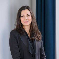 A person wearing a dark blazer stands indoors near a wall and dark curtains.