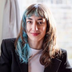 A person with long hair that includes a blue streak is wearing a dark jacket and light top, seated in front of a softly lit background.