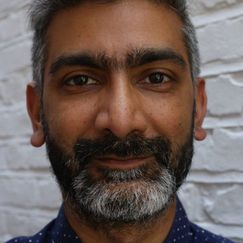 A person standing in front of a white brick wall, shown from the shoulders up, wearing a dark blue shirt with small white polka dots.