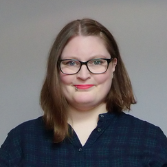 A person with shoulder‑length straight hair is shown from the torso up, wearing a dark, check‑patterned shirt against a plain, light‑colored background.