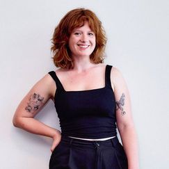 A professional photo of a woman dressed in black, with red hair and tattoos.