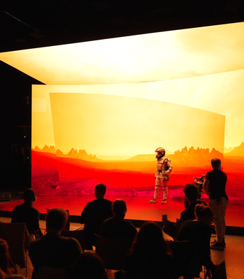The CoSTAR Screen Lab LED stage is lit up displaying a Martian landscape. An actor in a spacesuit is on the stage. A cameraman is shooting the scene from the side. A group of spectators watch from seats in front of the stage.