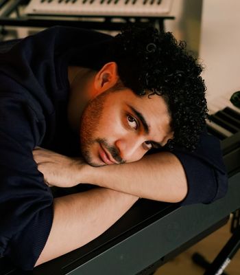 Portrait of a man with brown skin and dark hair leaning on a keyboard with his head on his forearm looking sideways at the camera