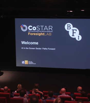 A speaker stands at a podium in a dark auditorium, addressing an audience seated in red chairs. A large screen behind the speaker displays the CoSTAR Foresight Lab and BFI logos with the title “Welcome – AI in the Screen Sector: Paths Forward.”