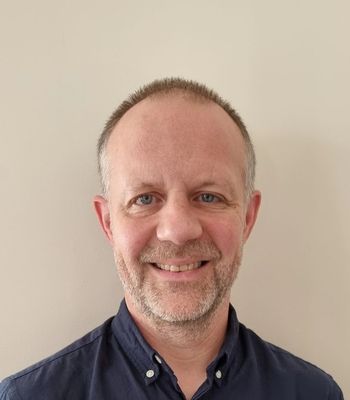 A person wearing a dark button‑up shirt stands against a plain light‑coloured wall.