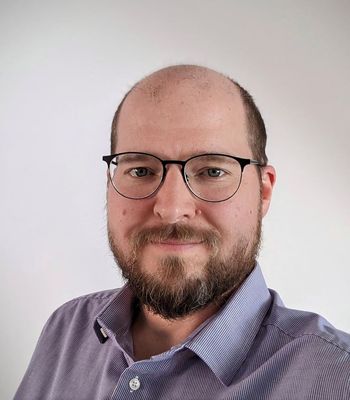 A person in a light purple collared shirt stands against a plain light background.