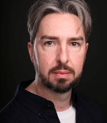 A person shown from the shoulders up, wearing a dark button-up shirt over a light-colored top, standing against a dark background.