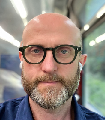 A person wearing glasses and earbuds, with bright ceiling lights and a window showing greenery outside.