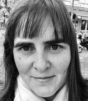 A person with medium-length hair stands outdoors in a black‑and‑white photo, with shops and parked scooters visible in the background.