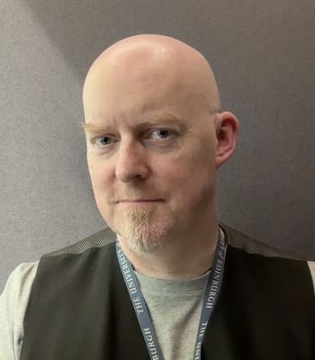 A person is shown from the shoulders up against a plain grey background, wearing a light-colored shirt, a dark vest, and a lanyard with text reading “THE UNIVERSITY OF EDINBURGH.”
