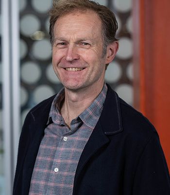 A person wearing a plaid shirt and a dark jacket stands indoors in front of a patterned background.