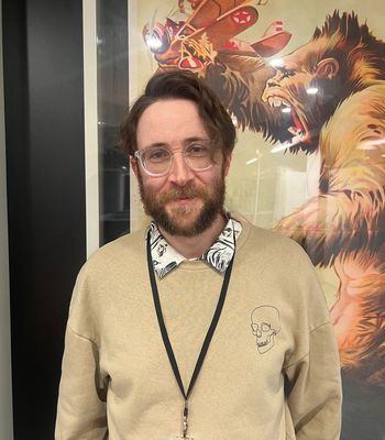 A person wearing a beige sweater with a skull design stands in front of a large poster featuring a stylized creature.