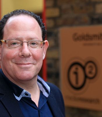 A person wearing a dark blazer over a collared shirt stands outdoors near a brick wall, with a sign in the background displaying the Goldsmiths University of London logo.