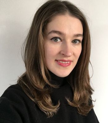 A person with long hair that includes a blue streak is wearing a dark jacket and light top, seated in front of a softly lit background.