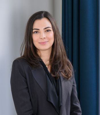 A person wearing a dark blazer stands indoors near a wall and dark curtains.