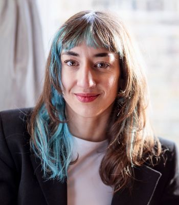 A person with long hair that includes a blue streak is wearing a dark jacket and light top, seated in front of a softly lit background.