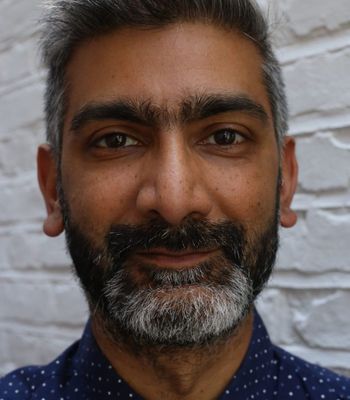 A person standing in front of a white brick wall, shown from the shoulders up, wearing a dark blue shirt with small white polka dots.