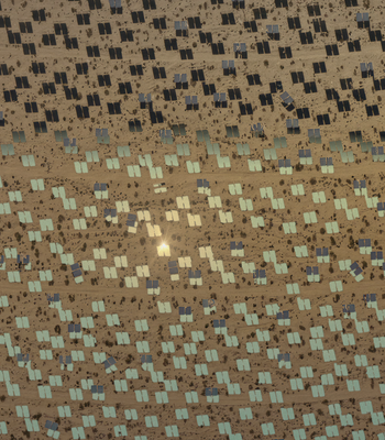 A film still from The Great Endeavour, directed and designed by Liam Young, 2024. The image shows an aerial view of a large solar farm in a desert landscape. Rows of solar panels are arranged in a grid pattern, with some sections appearing darker and others lighter, possibly due to the angle of sunlight or different types of panels. The ground is sandy and dotted with sparse vegetation. There is a bright reflection of sunlight near the centre of the image.