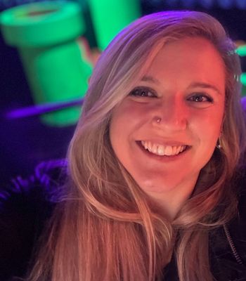 A person with long light-colored hair stands in a neon-lit environment with bright green and purple objects in the background.