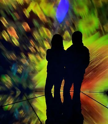 Two people stand in silhouette in front of a vivid, abstract light display with swirling green, yellow, blue, and red tones.