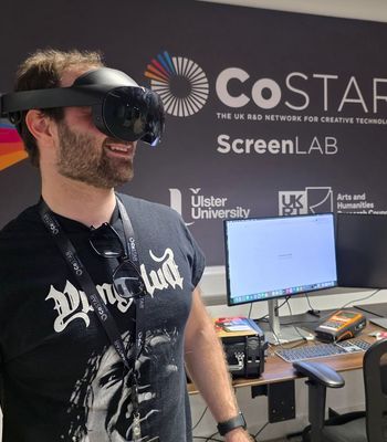 A workshop attendee wearing a mixed reality headset smiles while experiencing immersive technology in front of CoSTAR Screen Lab branding at Studio Ulster.