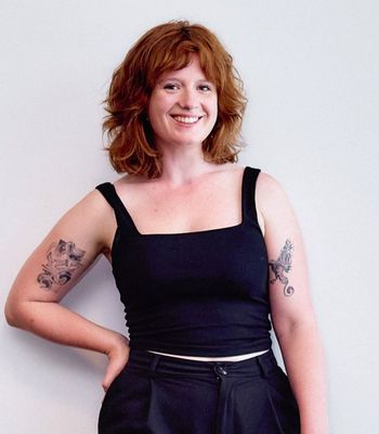 A professional photo of a woman dressed in black, with red hair and tattoos.
