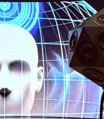 A multi-directional dodecahedron loudspeaker array stands in front of a digital display featuring a stylised human head and blue sound wave graphics, illustrating immersive spatial audio technology.