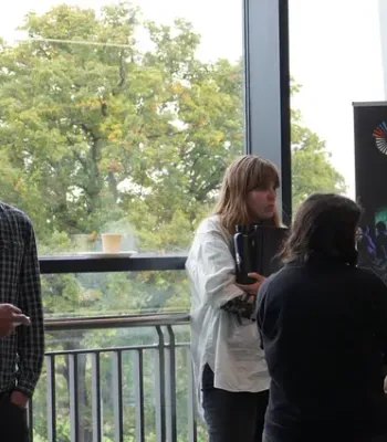 Five adults (two men and three women) are standing and chatting in a bright, modern room by a COSTAR promotional banner. Some are holding coffee cups, and trees are visible through large windows in the background.