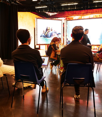 A group of people sits in blue chairs arranged in a circle inside a dim theater studio, facing a brightly lit stage with large framed digital art projections. One person uses a laptop while others listen and chat; faces are blurred for privacy.