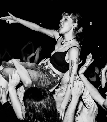 Black-and-white photo of a female crowd-surfing performer being carried above a packed concert crowd. She wears a black crop top and jeans, has tattoos on her left arm, and points outward with her right arm while fans reach up to support her