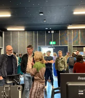 Interior of a conference/event space with a group of people mingling. In the foreground there are computer monitors; left wall banners display logos for Royal Holloway University of London and Surrey. The room has dark walls, bright ceiling lights, a blue floor, and an exit door with an illuminated green sign at the far end. Attendees are dressed casually and carry bags.