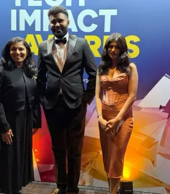 Three individuals posing at an awards event in front of a colorful backdrop that reads “Impact Awards.” From left to right: a woman in a black outfit, a man in a black tuxedo, and a woman in a brown dress.