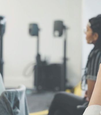 Close-up of a person with short hair and round glasses, resting their chin on their hand and holding a pen, in a classroom or workshop setting with blurred participants and equipment in the background.