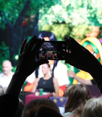 Silhouetted hands hold a smartphone up to photograph a panel discussion on a stage at a live event; the phone screen shows the panelists against a bright, leafy backdrop with audience heads in the foreground.