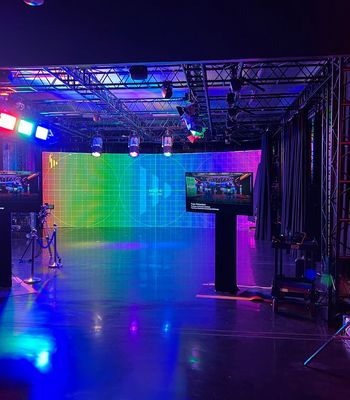 A stage setup featuring a rainbow-colored grid LED backdrop, metal truss lighting rig, and two display screens on tall stands. The glossy floor reflects blue and magenta lights, with various AV equipment and cables on the ground and a person seated at the left.